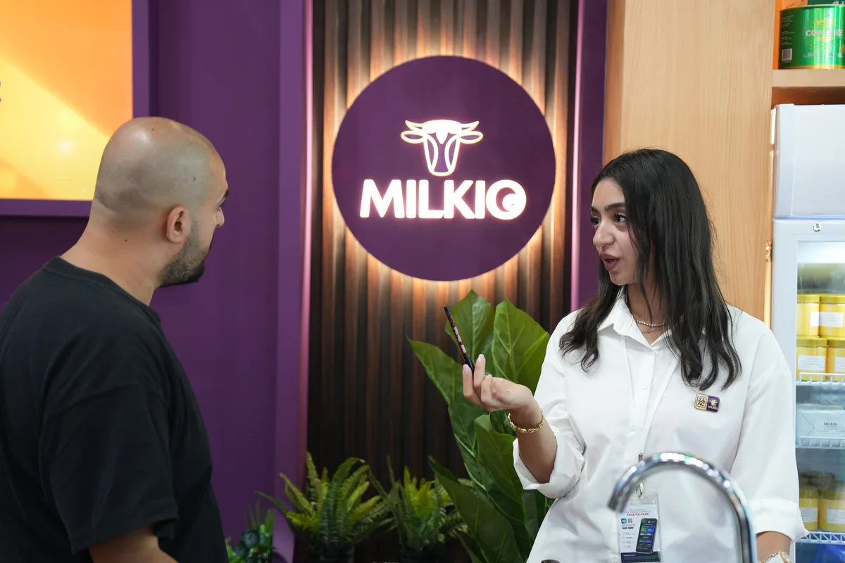 A ZAAC team member in a white shirt explaining the range to a visitor beside the Milkio sign